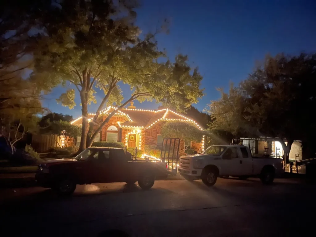 Barefoot crew trucks at dusk in front of a one-story home outlined in warm white C9 Christmas lights