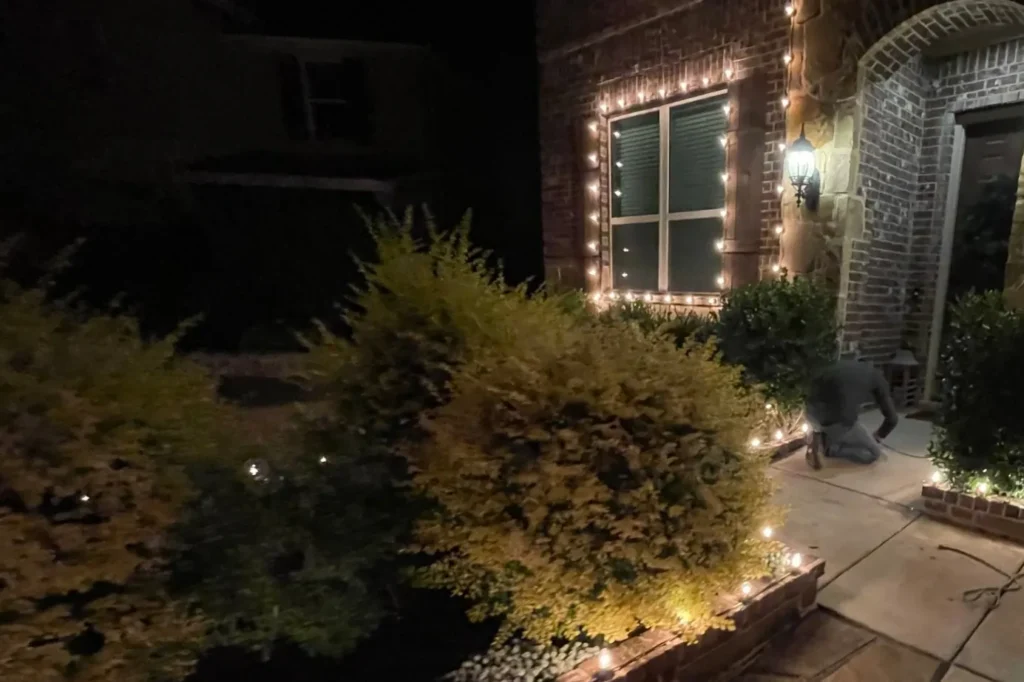 Barefoot crew member installing C9 Christmas lights along a front entry and flower bed on install day