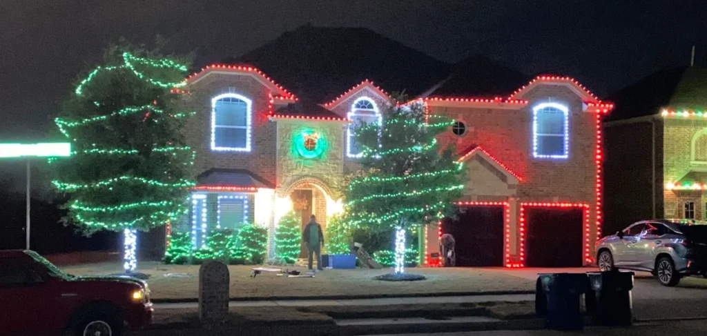 Full-color Christmas display with red roofline lights, white window outlines, and green-wrapped yard trees