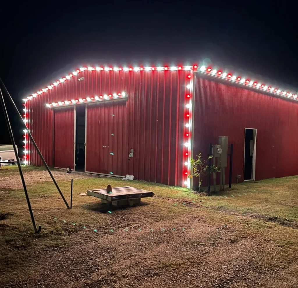 Red and white C9 Christmas lights outlining the roofline and doors of a red metal barn