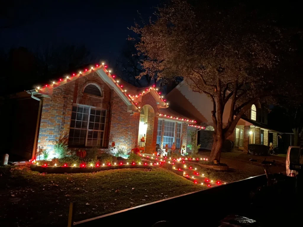 Red and white C9 Christmas lights along a one-story brick roofline with lit flower bed borders and front steps