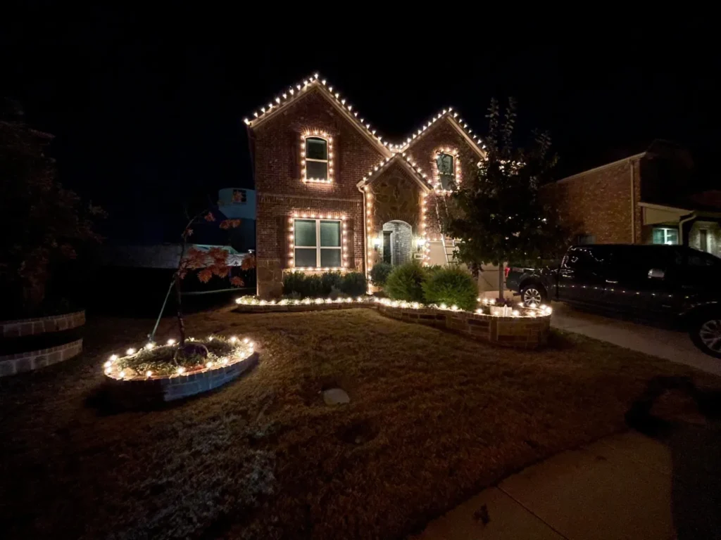 Warm white C9 Christmas lights outlining the roofline, windows, and flower beds of a two-story brick home in North Texas